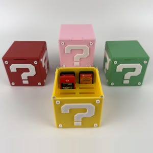 Könnte beinhalten: Vier würfelförmige Aufbewahrungsboxen für Spielkassetten mit einem weißen Fragezeichen-Design auf jeder Seite. Die roten, rosafarbenen und grünen Boxen sind geschlossen, während die gelbe Box geöffnet ist und zwei Spielkassetten im Inneren zeigt.