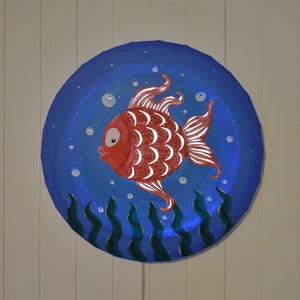 May include: A red fish with white scales and a blue background. The fish is surrounded by white bubbles and green seaweed. The fish is painted on a round paper plate.