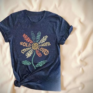 Navy blue T-shirt featuring colorful positive affirmation words such as brave, loved, strong, and courageous arranged in a heart-shaped design.