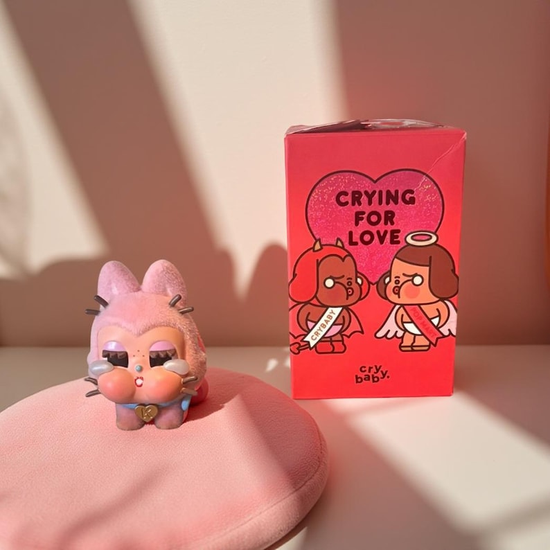 Pastel pink Crybaby collectible figurine with teardrop details, displayed beside a red “Crying for Love” box on a soft pink surface.