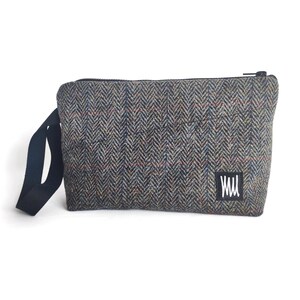 May include: A rectangular tweed crossbody bag with a black strap and zipper. The bag features a herringbone pattern in shades of gray, brown, blue, and red. A white label with black text is on the lower right corner.
