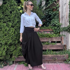 May include: A white long-sleeved button-down shirt with a gathered front detail paired with a high-waisted black maxi skirt featuring a draped side panel, a wide waistband, and a floor-length silhouette, worn with beige platform heels and sunglasses.