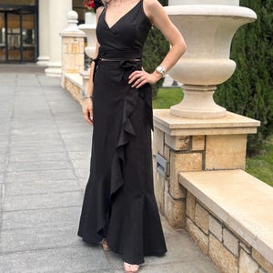 Puede incluir: Un conjunto negro de dos piezas que consta de un top cruzado y una falda larga. El top tiene cuello en V y se ata a la cintura. La falda presenta un detalle de volantes en el lateral. El conjunto se usa al aire libre.