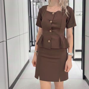 May include: A brown two-piece outfit featuring a short-sleeved top with wooden buttons and a peplum waist, paired with a matching skirt. The skirt has a front slit. The outfit is a solid color and appears to be made of a woven fabric.