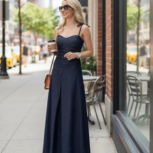 Peut inclure: Une femme porte un ensemble deux pièces en jean bleu foncé, composé d'un haut à fines bretelles et d'une longue jupe. Elle tient une tasse de café et un sac à main marron. Elle porte des lunettes de soleil et une montre.