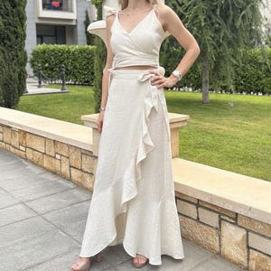 Puede incluir: Un conjunto de dos piezas color crema, con un top cruzado con detalles de lazo y una falda cruzada a juego con un borde con volantes. El conjunto está hecho de una tela ligera, perfecto para el clima cálido. La falda llega hasta los tobillos.