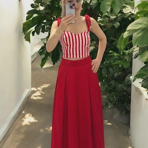 Puede incluir: Una falda roja y un top a rayas rojas y blancas. El top tiene tirantes anudados y un escote corazón. La falda es de color rojo liso y es larga hasta el suelo. El atuendo se usa en un área bien iluminada con plantas verdes en el fondo.