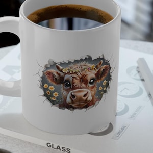 Könnte beinhalten: Weiße Keramiktasse mit einer Illustration eines braunen und weißen Kuhkopfes. Die Kuh trägt eine Blumenkrone und schaut aus einem Loch in der Tasse. Die Tasse ist mit Kaffee gefüllt.