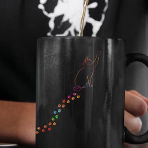 Puede incluir: Una taza de café negra con un rastro de huellas de patas de arcoíris y una silueta de gato negro. La taza está llena de café y se está vertiendo desde una tetera negra moteada.