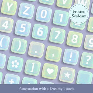Puede incluir: Un conjunto de fichas de alfabeto azul pastel y verde iridiscente con letras blancas y signos de puntuaci&oacute;n. Las fichas est&aacute;n dispuestas en una cuadr&iacute;cula sobre un fondo morado claro. El texto "Frosted Seafoam" est&aacute; escrito en un c&iacute;rculo blanco en la esquina superior derecha de la imagen. El texto "Punctuation with a Dreamy Touch." est&aacute; escrito en blanco en la parte inferior de la imagen.
