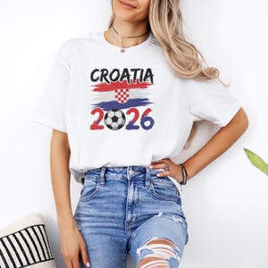 A woman wearing a white t-shirt with a graphic design of the Croatia national flag and the year "2026" printed on it, standing next to a potted plant.