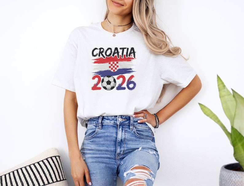 A woman wearing a white t-shirt with a graphic design of the Croatia national flag and the year "2026" printed on it, standing next to a potted plant.