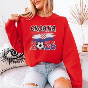 A woman wearing a red sweater with the text "Croatia 2026" and a soccer ball graphic, sitting on a bed with a decorative pillow.