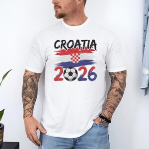 A man wearing a white t-shirt with the text "Cromia 2026" and a graphic of the Croatia national flag, standing in front of a plant and a denim jacket.