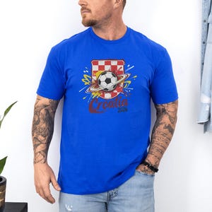 A man wearing a blue t-shirt with a graphic of a soccer ball and a red and white checkered flag, standing against a white wall.
