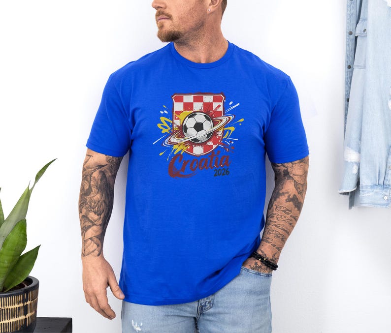 A man wearing a blue t-shirt with a graphic of a soccer ball and a red and white checkered flag, standing against a white wall.