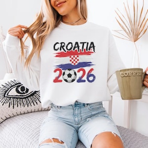 A woman wearing a white sweatshirt with a graphic of the Croatia national flag and the year "2026" printed on it, sitting on a bed and holding a coffee mug.