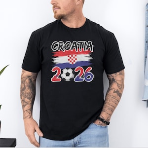 A man wearing a black t-shirt with the text "CROATIA 2026" printed on it, standing in front of a plant and a denim jacket.