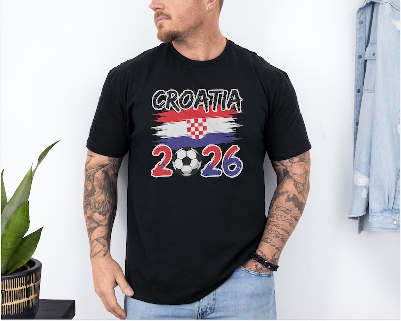 A man wearing a black t-shirt with the text "CROATIA 2026" printed on it, standing in front of a plant and a denim jacket.