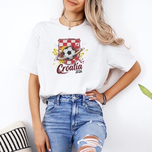 A woman wearing a white t-shirt with a graphic of a soccer ball and the text "Croatia 2006" stands next to a potted plant.