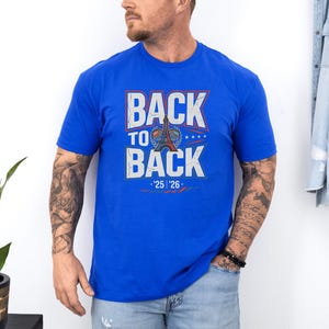 A man wearing a blue t-shirt with the text "BACK TO BACK" and "25 26" printed on it, standing against a white wall.