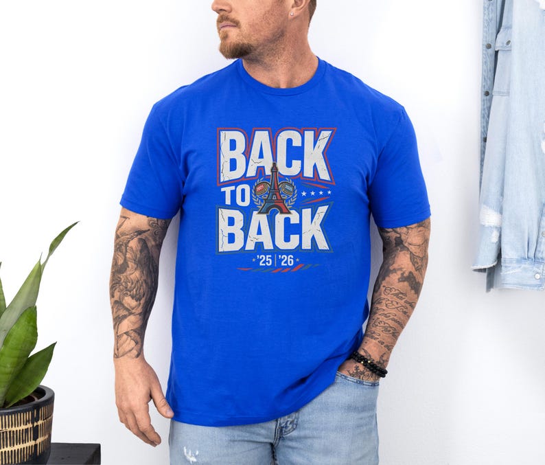 A man wearing a blue t-shirt with the text "BACK TO BACK" and "25 26" printed on it, standing against a white wall.