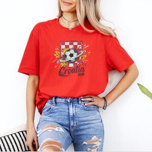 A woman wearing a red t-shirt with a graphic of a soccer ball and a red and white striped shield, standing in front of a white wall.