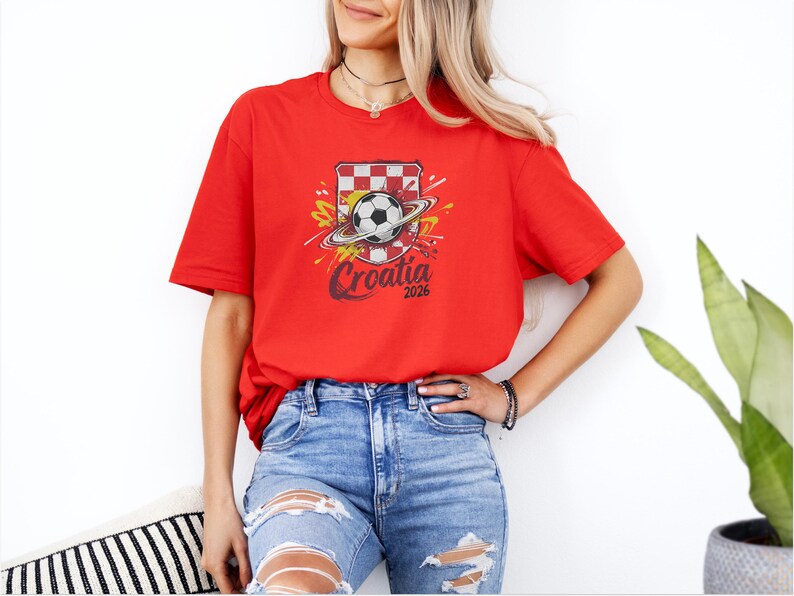 A woman wearing a red t-shirt with a graphic of a soccer ball and a red and white striped shield, standing in front of a white wall.