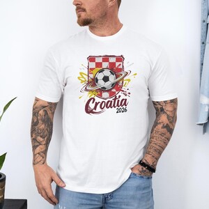 A man wearing a white t-shirt with a graphic of a soccer ball and a red and white checkered flag, along with the text "Croatia 2026".