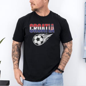 A man wearing a black t-shirt with the word "Croatia" printed on it, standing in front of a plant and a denim jacket.