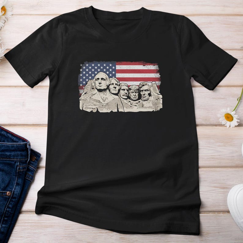 A black t-shirt with a graphic design featuring the Statue of Liberty and the faces of three other figures, all set against an American flag.