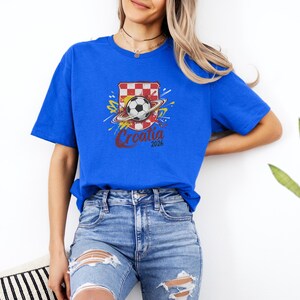 A woman wearing a blue t-shirt with a graphic of a soccer ball and a red and white shield, standing against a white wall.