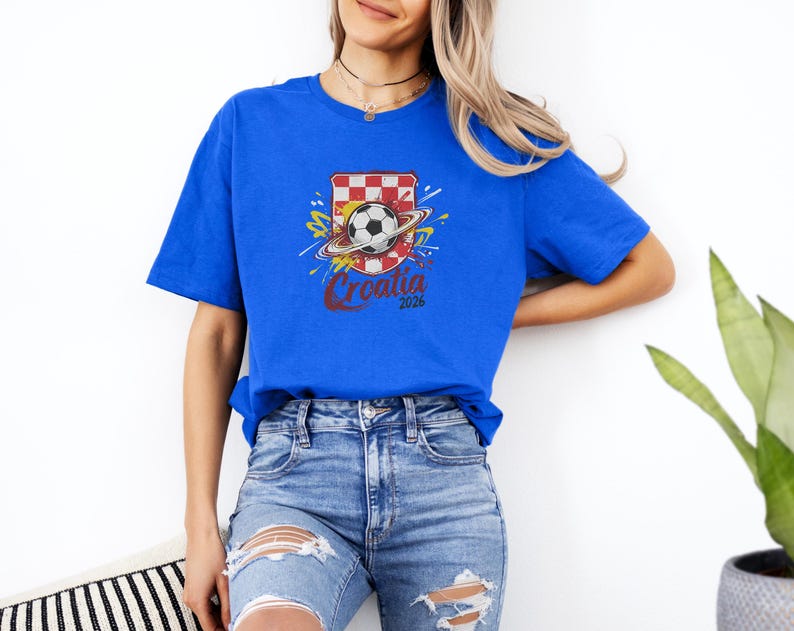 A woman wearing a blue t-shirt with a graphic of a soccer ball and a red and white shield, standing against a white wall.