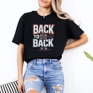 A woman wearing a black t-shirt with the text "BACK TO BACK" and jeans, standing against a white wall with a potted plant in the background.