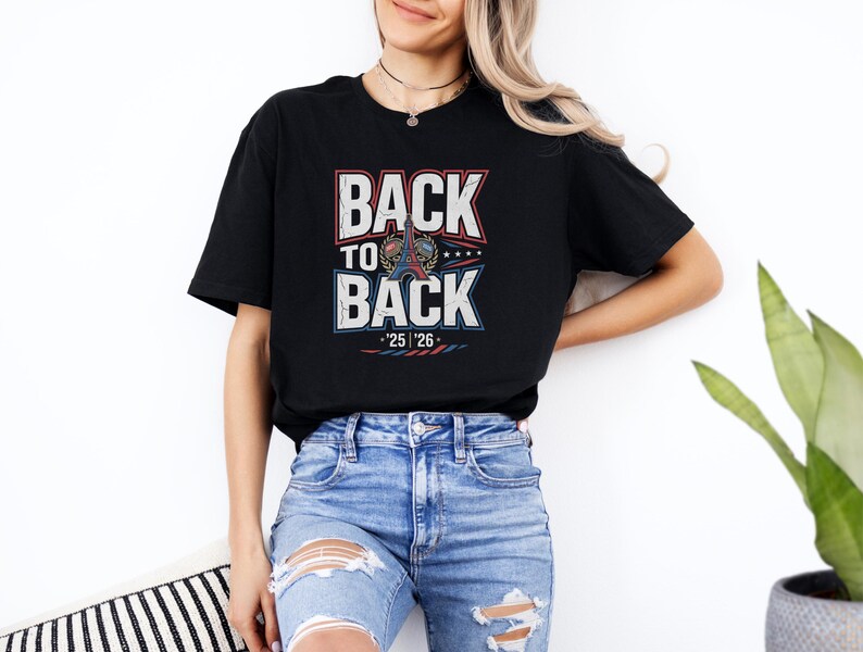 A woman wearing a black t-shirt with the text "BACK TO BACK" and jeans, standing against a white wall with a potted plant in the background.