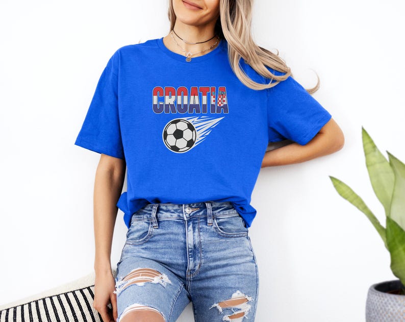 A woman wearing a blue t-shirt with the word "Croatia" and a soccer ball graphic, paired with distressed jeans, stands against a white wall.