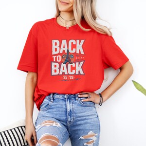 A woman wearing a red t-shirt with the text "BACK TO BACK" and jeans stands in front of a white wall, with a potted plant visible to her right.