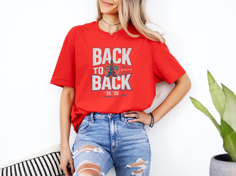 A woman wearing a red t-shirt with the text "BACK TO BACK" and jeans stands in front of a white wall, with a potted plant visible to her right.