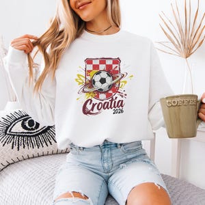 A woman wearing a white sweater with a graphic of the Croatia national soccer team and holding a coffee mug is sitting on a bed.