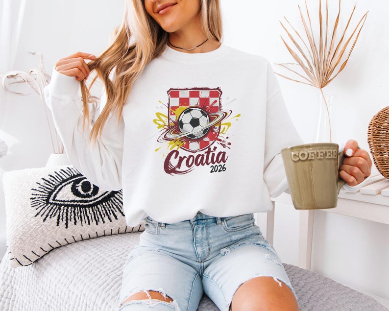 A woman wearing a white sweater with a graphic of the Croatia national soccer team and holding a coffee mug is sitting on a bed.