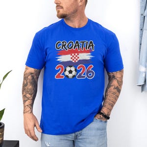 A man wearing a blue t-shirt with the text "Croatia 2026" and a soccer ball graphic stands next to a plant.