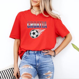 A woman wearing a red t-shirt with the word "Croatia" and a graphic of a soccer ball, paired with distressed jeans, stands in front of a white wall.