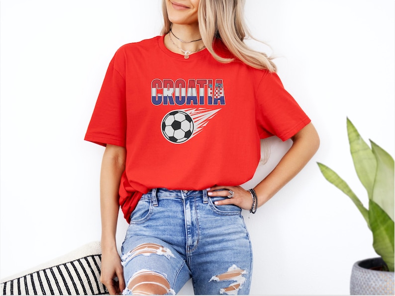 A woman wearing a red t-shirt with the word "Croatia" and a graphic of a soccer ball, paired with distressed jeans, stands in front of a white wall.
