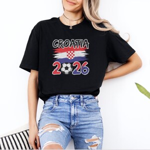 A woman wearing a black t-shirt with the text "GROATIA 2026" and a graphic of the Croatia national flag, along with distressed jeans, stands in front of a white wall.