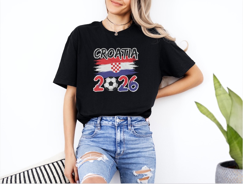A woman wearing a black t-shirt with the text "GROATIA 2026" and a graphic of the Croatia national flag, along with distressed jeans, stands in front of a white wall.
