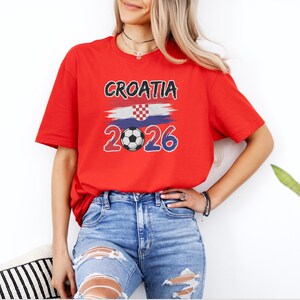 A woman wearing a red t-shirt with the text "CROATIA 2026" and distressed jeans stands in front of a white wall, with a potted plant visible to her right.