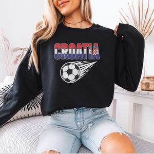 A woman wearing a black sweatshirt with the word "Croatia" printed on it, sitting on a bed with a soccer ball design.
