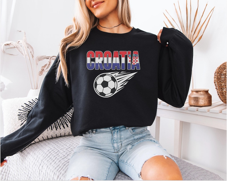 A woman wearing a black sweatshirt with the word "Croatia" printed on it, sitting on a bed with a soccer ball design.