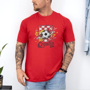 A man wearing a red t-shirt with a soccer ball and a red and white checkered flag design, standing against a white wall.