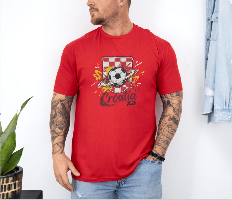 A man wearing a red t-shirt with a soccer ball and a red and white checkered flag design, standing against a white wall.